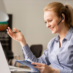 A smiling woman with a headset gestures during a video call, holding a clipboard in front of her laptop. This scene illustrates a professional using Microsoft Teams compliance recording or VoIP call recording as part of a cloud-based communication solution.